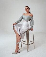 Elegant woman in pastel green top, silver skirt and high heels sitting at high metal chair with crossed legs. Stunning gorgeous fashion female model with natural make-up and pony tail