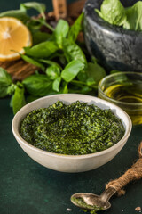  Pesto sauce in bowl with ingredients on rustic green table. Traditional Italian pesto recipe for making fettuccine, pasta, bruschetta.