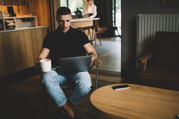 Young tattooed man working on laptop and drinking coffee in kitchen