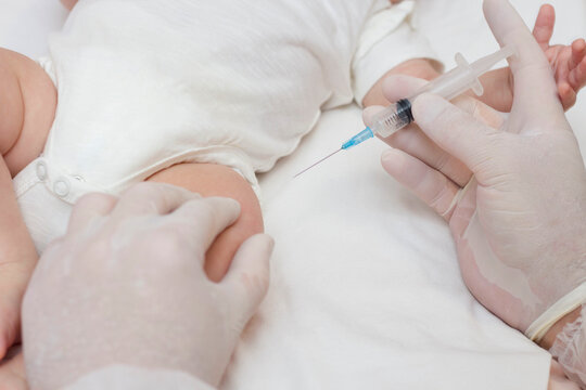 The doctor's hands in rubber protective gloves hold a syringe. An infant receiving the vaccine in the leg. Visit to the pediatrician in the hospital. A two-month-old baby. To close.