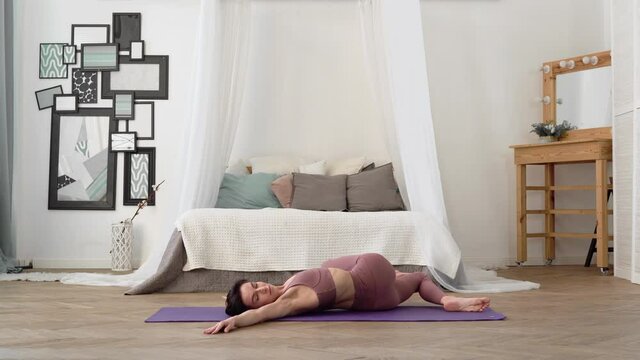 Slender, flexible, caucasian middle-aged woman with black hair in beige tracksuit performs standing asanas Supine Spinal Twist Supta Matsyendrasana on purple yoga mat in bright room with bed, window