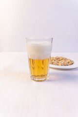 Glass of light beer with foam and plate with pistaches on white background