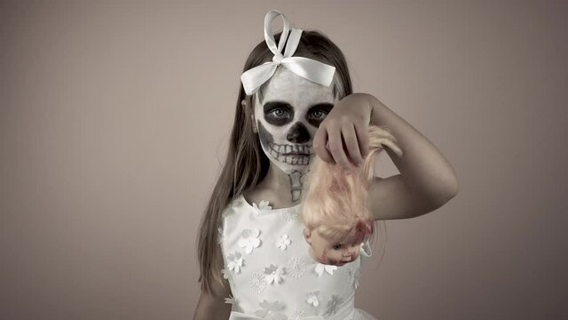A little girl in a dress with a painted face raises her hand with the bloody head of a doll.