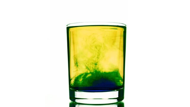 A moving liquid blue pigment solute at yellow colored water to create green solution in shaking glass placed on a pure white background. Majestic patterns under trembling water surface