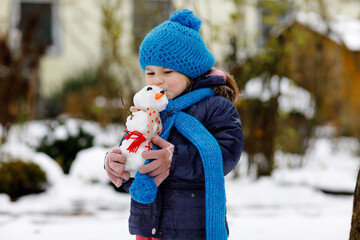 Cute little toddler girl making mini snowman and eating carrot nose. Adorable healthy happy child...