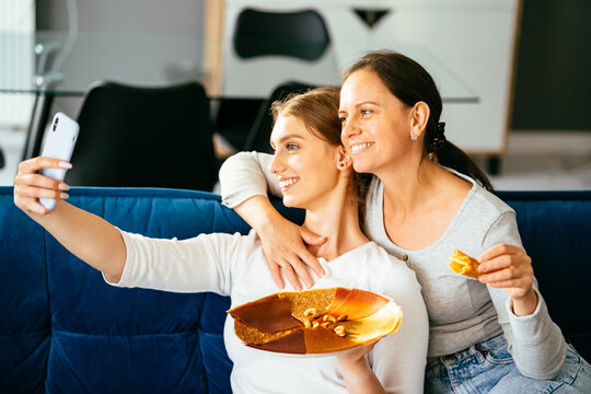 Happy women doing selphie by the smart phone. Mother and grown daughter sitting on the sofa at home, snuggled. They taking selphie, smiling eating berry pastille. Relationships and shared time.