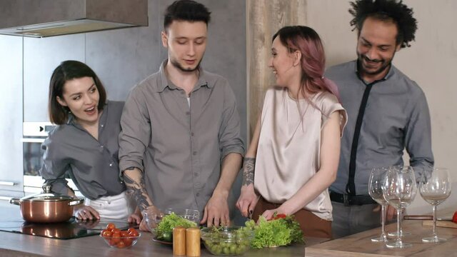 Cheerful diverse friends gathering for dinner party: couple making salad together in the kitchen and greeting man and woman walking to them and offering help