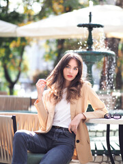 A woman on a summer terrace next to a fountain sits in a jacket and jeans