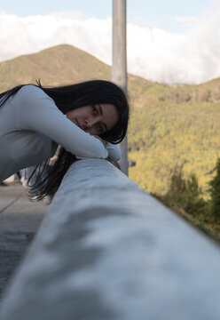 1 chica cauc&aacute;sica, vestida de blanco y de pelo negro recostada en un barandal al aire libre 