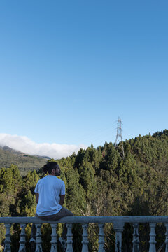 1 chico blanco de pelo largo y barba sentado en una baranda con fondo natural, torre el&eacute;ctrica y monta&ntilde;oso mirando al horizonte 