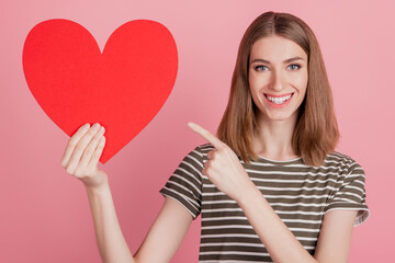 Portrait of reliable lady demonstrate red paper heart direct finger on pink background