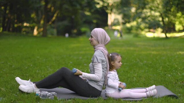Family sport activity outdoors. Pretty sporty fit Muslim Arabian woman working out with dumbbells together with her cute 4 years old daughter, sitting on yoga mat in the summer park