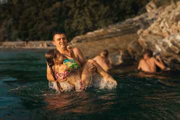 A young father holds his smiling daughter in his arms in the water of a mountain river. A family with one parent on vacation on the river on a sunny day at sunset.