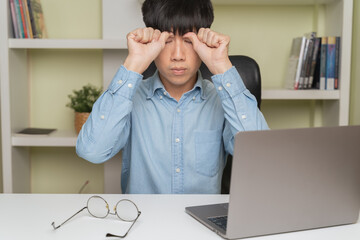 Tired young Asian businessman, man massaging close eyelids to reduce office syndrome, yoga to eyes sight relaxation for eyestrain after overworked at home workplace. Massaging office syndrome concept.