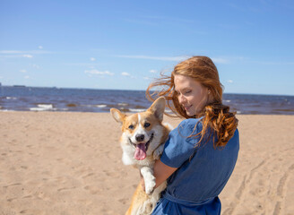 A girl with long red hair hugs her corgi dog tightly, on the beach, in the morning. Summer, friendship, beloved pet