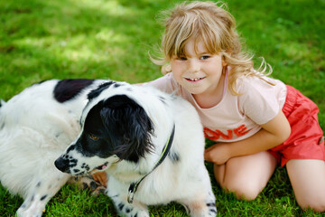 Naklejka na ściany i meble Cute little preschool girl playing with family dog in garden. Happy smiling child having fun with dog, hugging. Happy family outdoors. Friendship and love between animal and kids