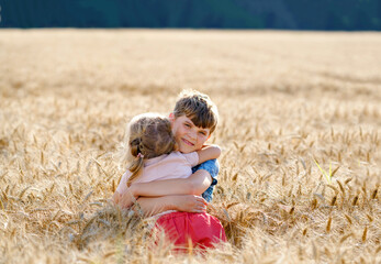 School kid boy and little sister, preschool girl hugging on wheat field. Two happy children playing together and having fun sunny summer day. Siblings in love.