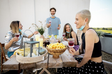 Young stylish friends in face masks have a festive dinner, toasting and having great summer time...