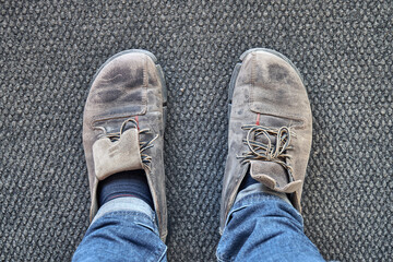 Legs of man in blue jeans and old brown shoes stand on gray carpet in workshop first point view