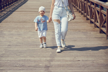 Adorable little kid boy in white hat walks with young mommy joining hands along stylish wooden footbridge on sunny summer day