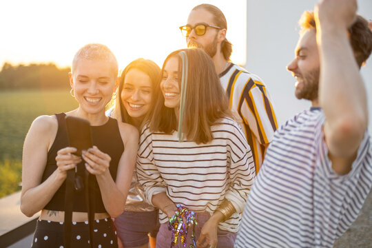 Young stylish friends have fun, showing something on phone and laugh outdoors. Hipsters, millennials hang out at rooftop terrace on sunset - Powered by Adobe