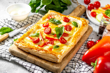 Puff pastry pie with mixed vegetables and mozzarella cheese on a serving board on a culinary table closeup. Delicious hearty pastries with tomatoes, zucchini, sweet pepper, spices and basil