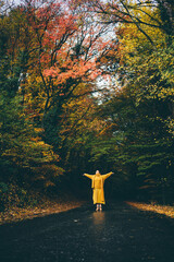 Cheerful young woman in stylish yellow clothes dancing along empty road across picturesque autumn forest with old colorful trees