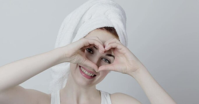 Beautiful young woman after shower with towel on her head, dancing and looking at camera with happy smile through the heart gesture formed with her hands