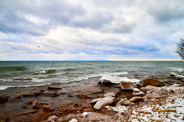 Nature landscape with dramatic sky and clouds, lake with ice, shore with snow and dark trees in...