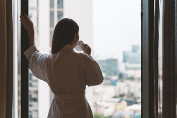 Relaxed Asian woman wearing bath robe drinking coffee in hotel room in morning 