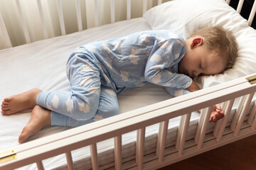 top view Cute little 2-3 years preschool baby boy kid sleeping sweetly in white crib during lunch rest time in blue pajama with pillow at home. Childhood, leisure, comfort, medicine, health concept
