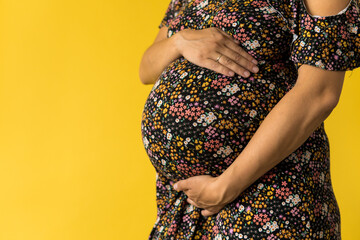 Motherhood, femininity, love, care, waiting, hot summer - bright croped Close-up unrecognizable pregnant woman in floral black dress with hands over tummy rub belly on yellow background, copy space
