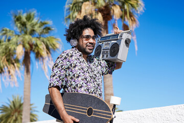 Young african american skater man listening music from vintage boombox stereo on the beach - obrazy, fototapety, plakaty