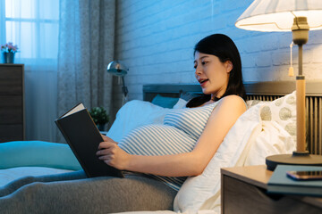 side portrait happy asian expectant mom sitting on bed is reading a book to her future baby by the...