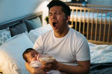 tired asian first time dad is yawning while sitting at bedside feeding his newborn girl bottled...