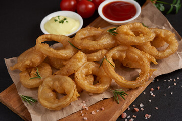 Homemade baked onion rings fries with mayonnaise, Tomato sauce and rosemary on wooden board. fast food products : onion rings on cutting board, on black stone background, unhealthy food.