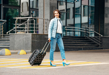 A satisfied woman walks down the street with a suitcase. Opening of borders between countries. The woman travels freely. Fall casual wear: jacket, sweater and jeans with blue high heels.