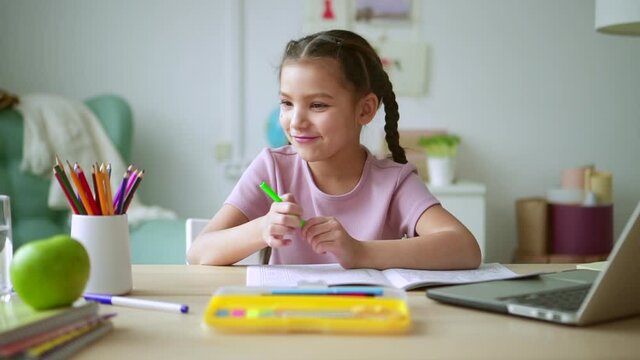 Online primary school education. Spbd Pretty brunette schoolgirl changes marker doing classwork during video lesson via modern laptop at desk in light room