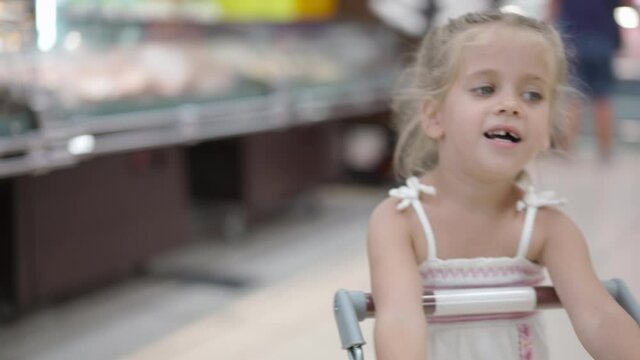 Handheld Effect Funny Cute Child Pushing Shopping Cart In Supermarket. Little Girl Buying With Special Shopping Cart For Children Kid Grocery Shopping. Adorable Baby Kid With Trolley 