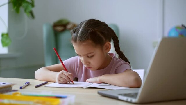 Online education at home. Spbd Serious brunette little girl pupil draws image in notebook with red pencil at videochat via laptop at table in light room