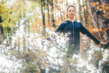 Fototapeta na wymiar Pretty tranquil woman with ponytail in blue contemporary tracksuit meditates among colorful autumn forest on nice sunny day
