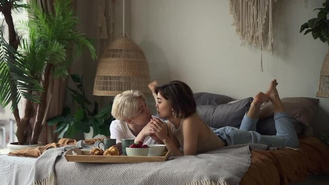 Lesbian couple at breakfast in bedroom. Spbd Happy young girlfriends hug and kiss spending time in modern bedroom at home in lazy morning