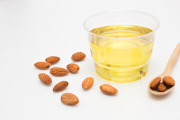 Yellow almond oil in a clear bowl with a wooden spoon next to it and several almonds on a white background