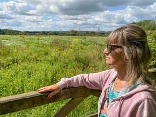 A lady in a pink hoody wearing sunglasses leans with hand on a wooden farm gate looking out towards a wild meadow in background.Sunny day with blue sky and white clouds - obrazy, fototapety, plakaty