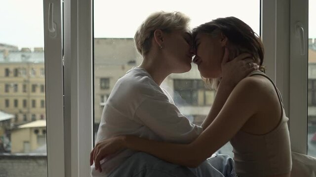 Two lesbian women kissing and hugging while sitting by window in apartment room spbd. Side view of beautiful young females embrace and kiss sensually, look with smiles and sit in light interior