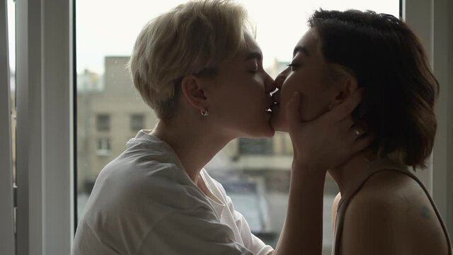 Lesbian couple love affection. Spbd Languid blonde lady kisses beloved Asian brunette girlfriend being intimate by window in light hotel room side view