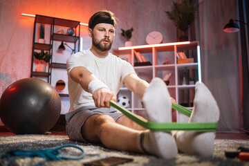 Bearded man with serious facial expression having evening workout with rubber band. Caucasian male person sitting on floor and doing exercises for arms.