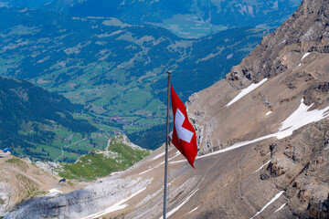 In the foreground the flag of Switzerland, in the background the icy mountains and the green valley 