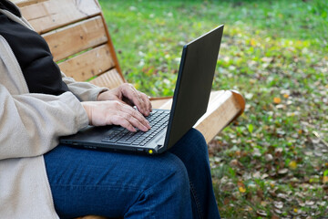 Пожилая женщина в джинсах работает на ноутбуке сидя на скамейке на открытом воздухе. Концепт удаленная работа, фриланс, заработок в интернет