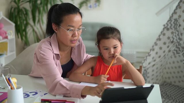 Home education with tutor. Spbd Asian woman former teacher with glasses helps little schoolgirl to do homework with tablet at table in light room